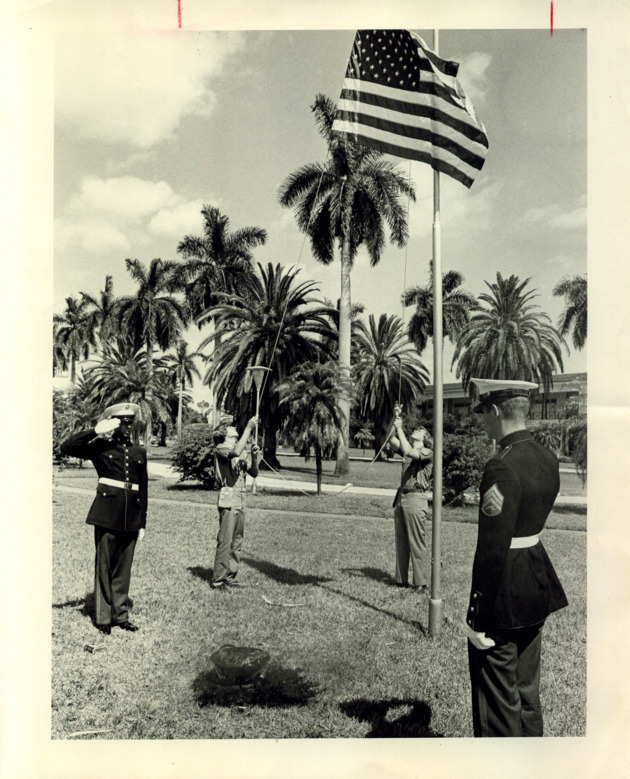 Two Marines in dress uniform salute as two others raise the flag in front of Adrian Hall